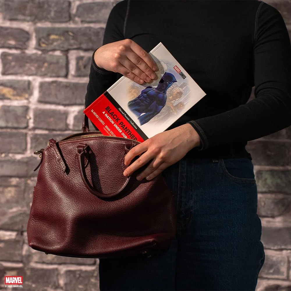 A person in black and jeans puts the book into a dark red leather handbag against a brick wall.